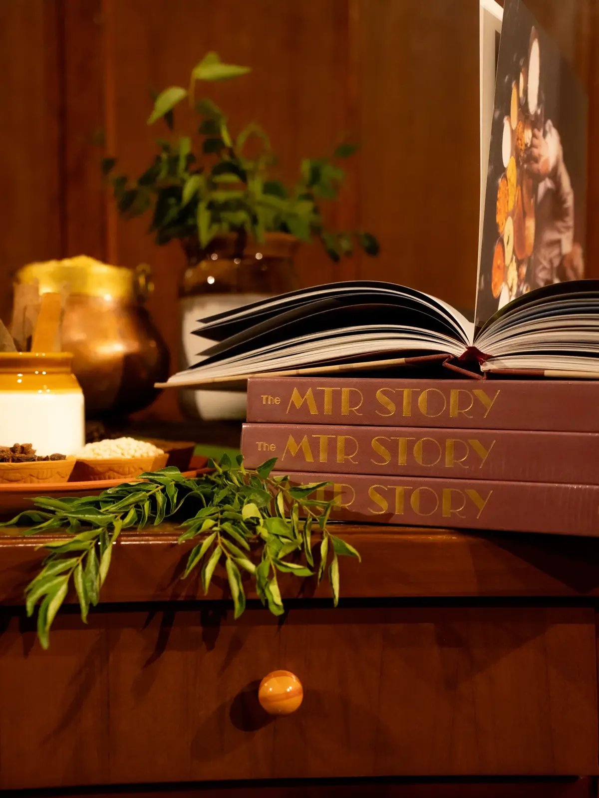 Stacked copies of The MTR Story with an open book, styled with curry leaves, spices, and brass vessels